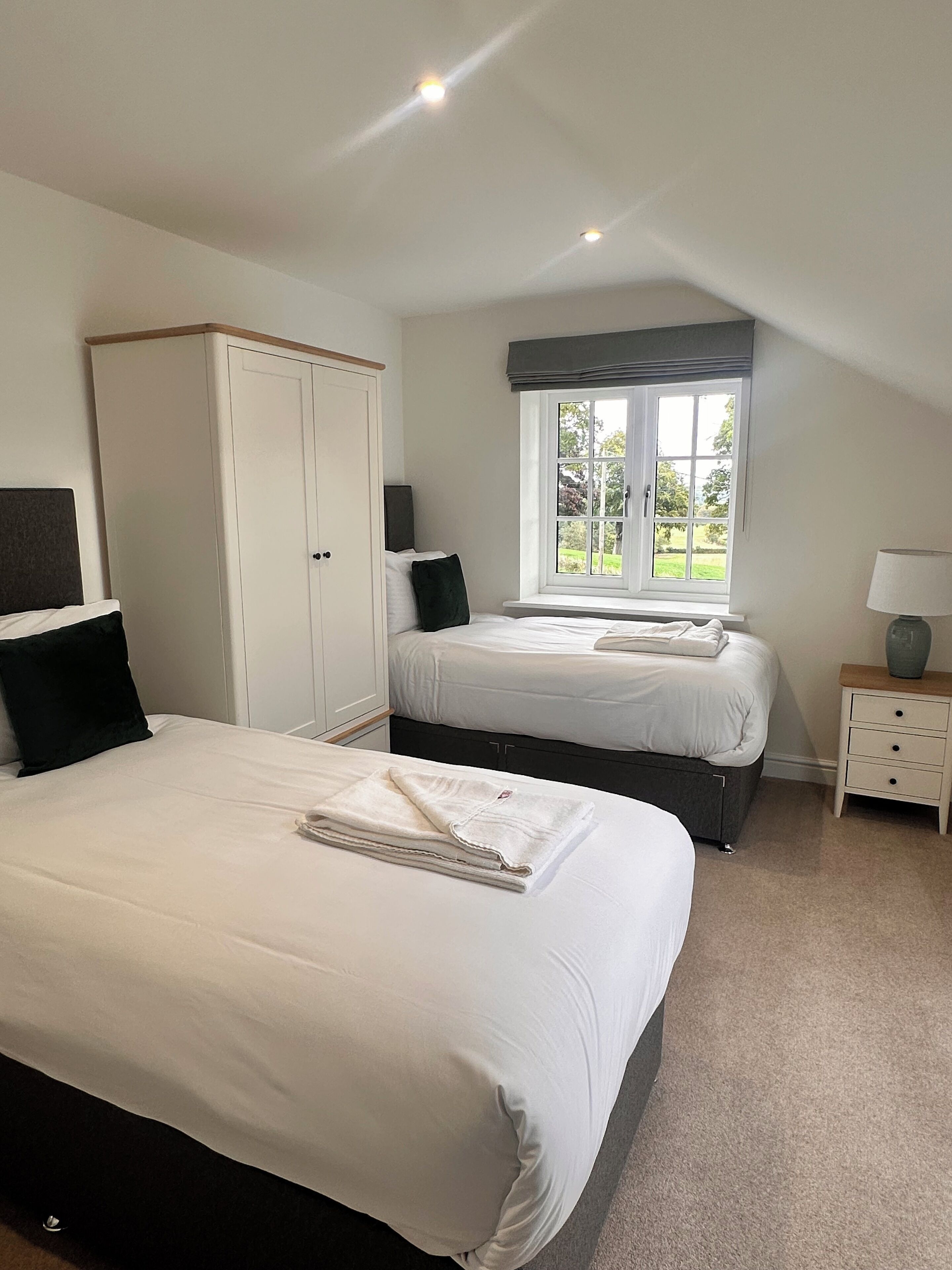 A bedroom with two beds, a wardrobe, a window with a view, and a bedside table with a lamp.