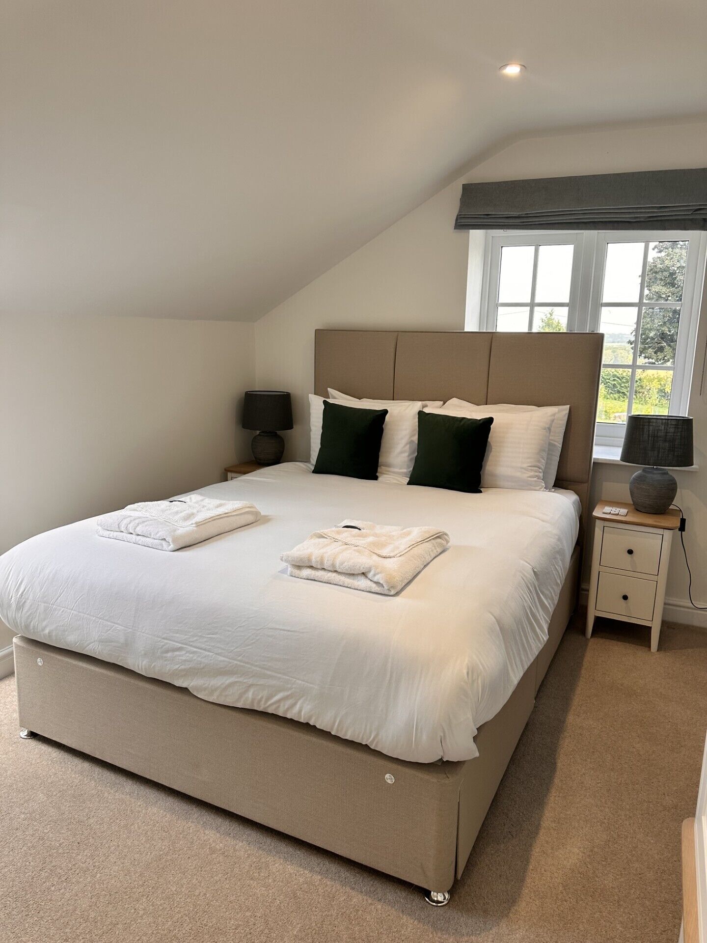 A neatly made bed with white linens and green pillows, a bedside table with a lamp, and a window with a view of greenery.