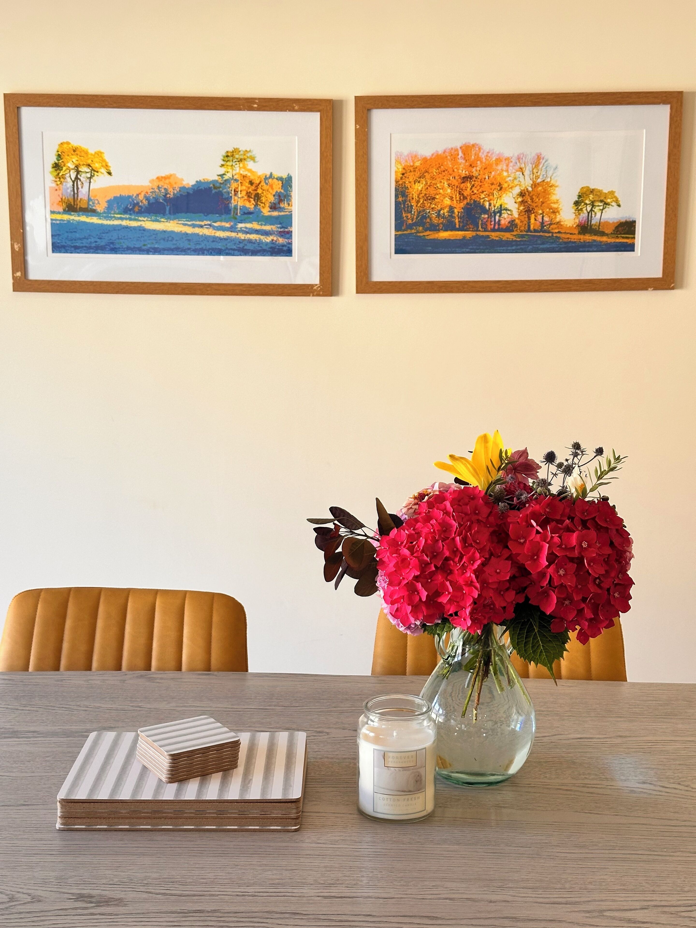 A dining table with a vase of flowers, a candle, and a stack of books. Two framed paintings are hung on the wall above.
