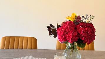 A dining table with a vase of flowers, a candle, and a stack of books. Two framed paintings are hung on the wall above.