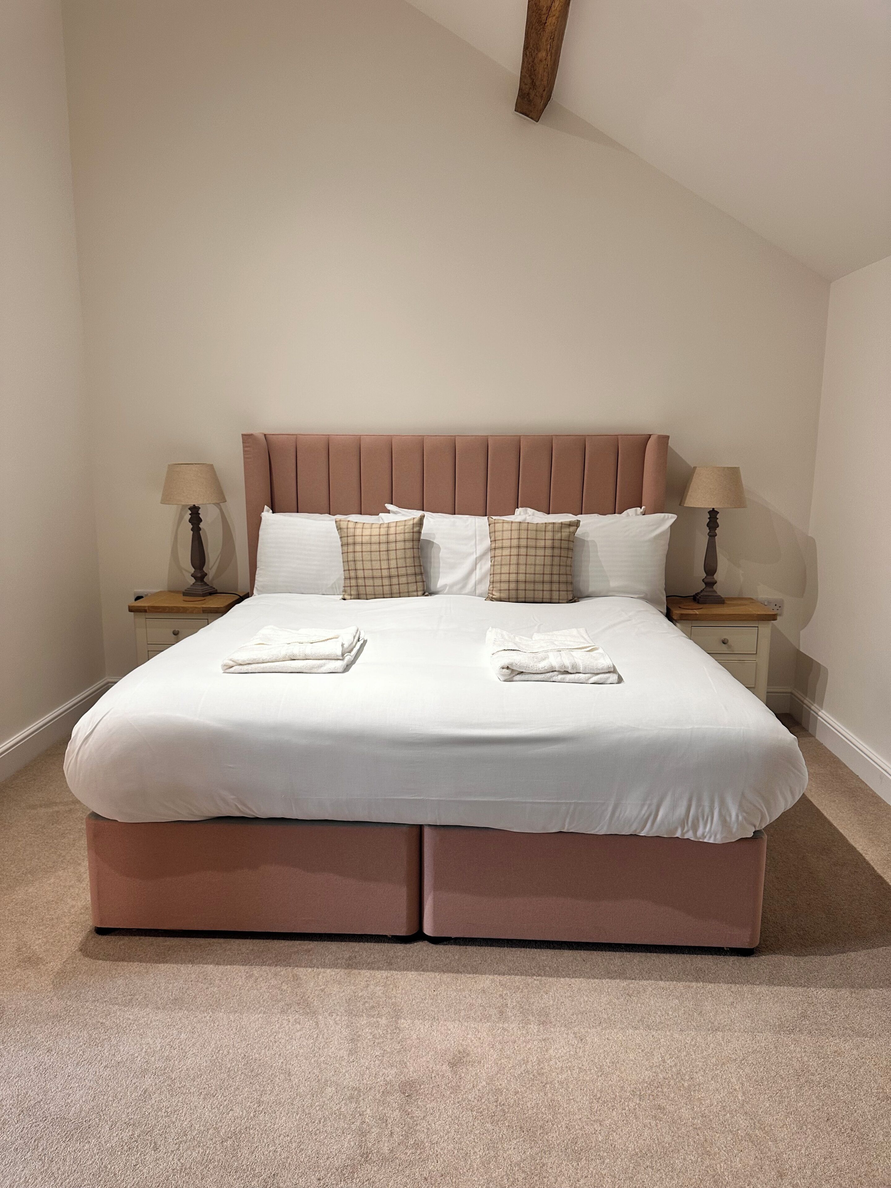 A neatly made bed with a pink headboard, two bedside tables with lamps, and a carpeted floor.