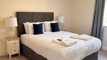 Una cama bien hecha con sábanas blancas y almohadas oscuras, una mesita de noche con una lámpara y una ventana con cortinas.