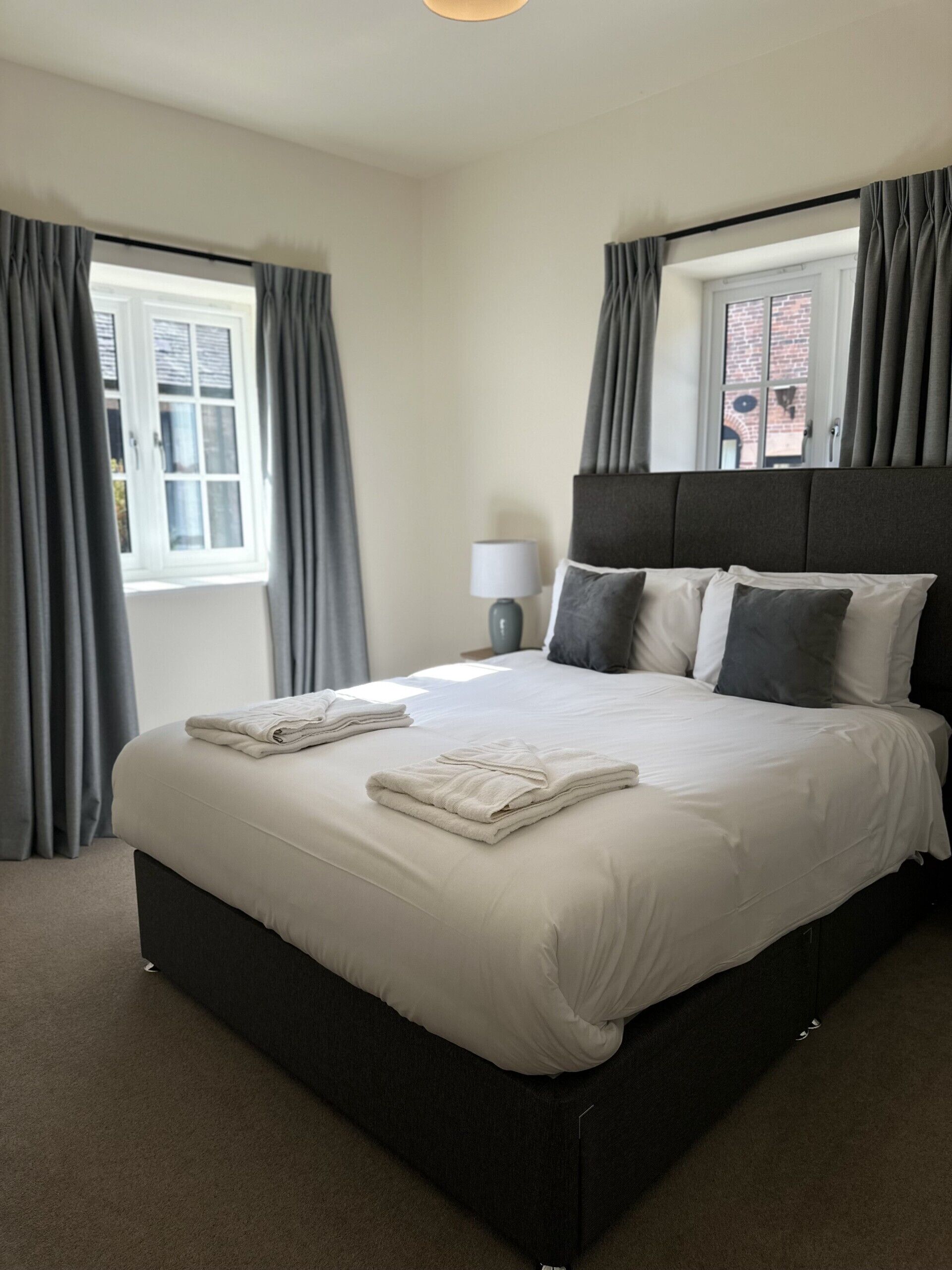 A neatly made bed with pillows and folded towels, a bedside lamp, and a window with curtains.