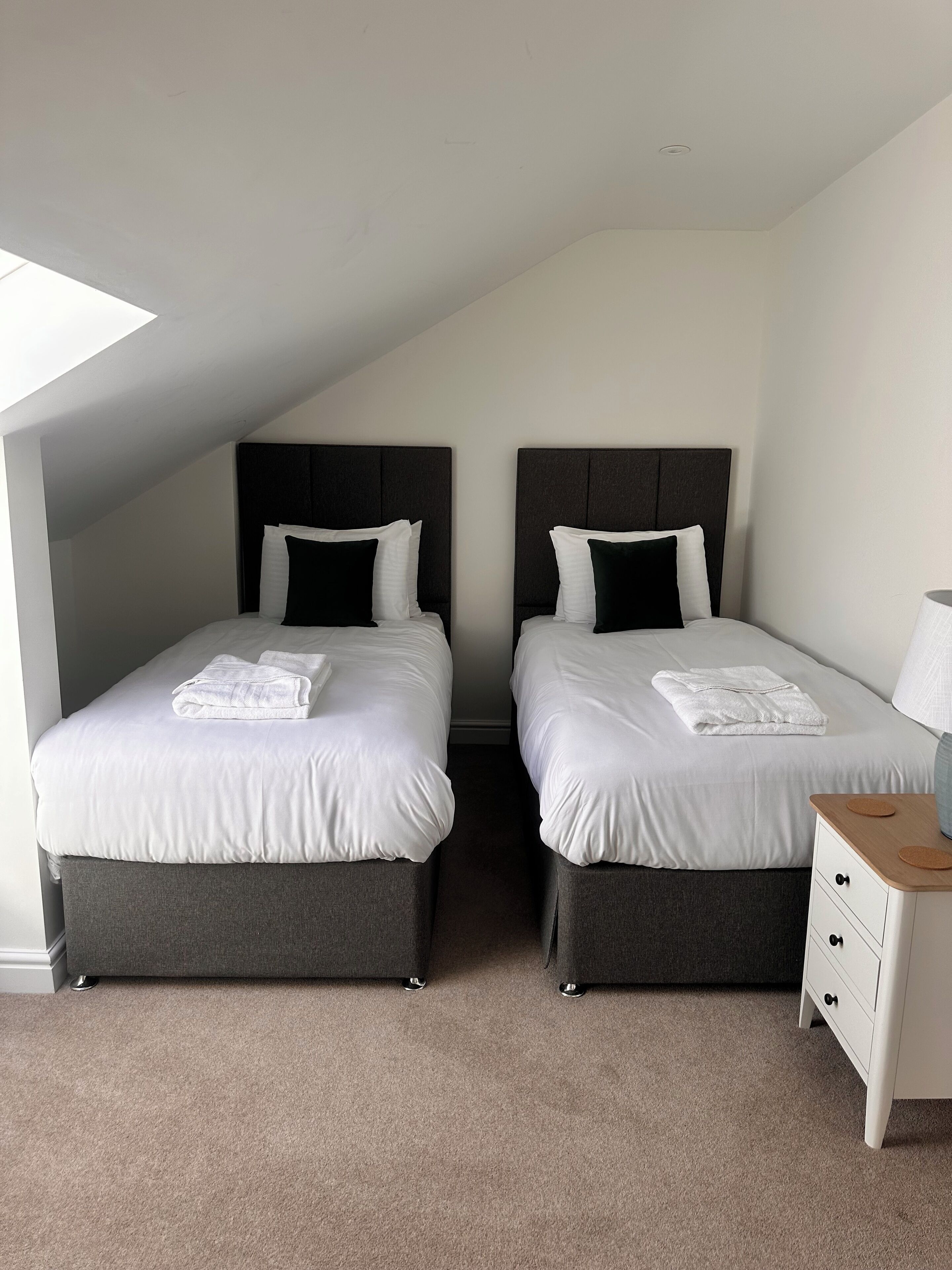 Ein Zimmer mit zwei Betten, jeweils mit dunklem Kopfteil und weißer Bettwäsche. Ein kleiner Nachttisch mit Lampe und Schublade steht bereit. Das Zimmer verfügt über eine geneigte Decke und einen Teppichboden.