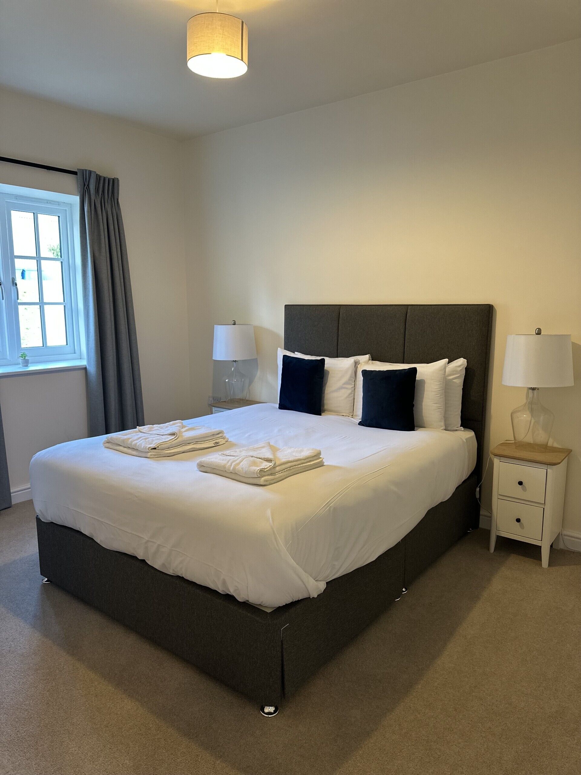A neatly made bed with white linens and folded towels, a bedside table with a lamp, and a window with curtains.