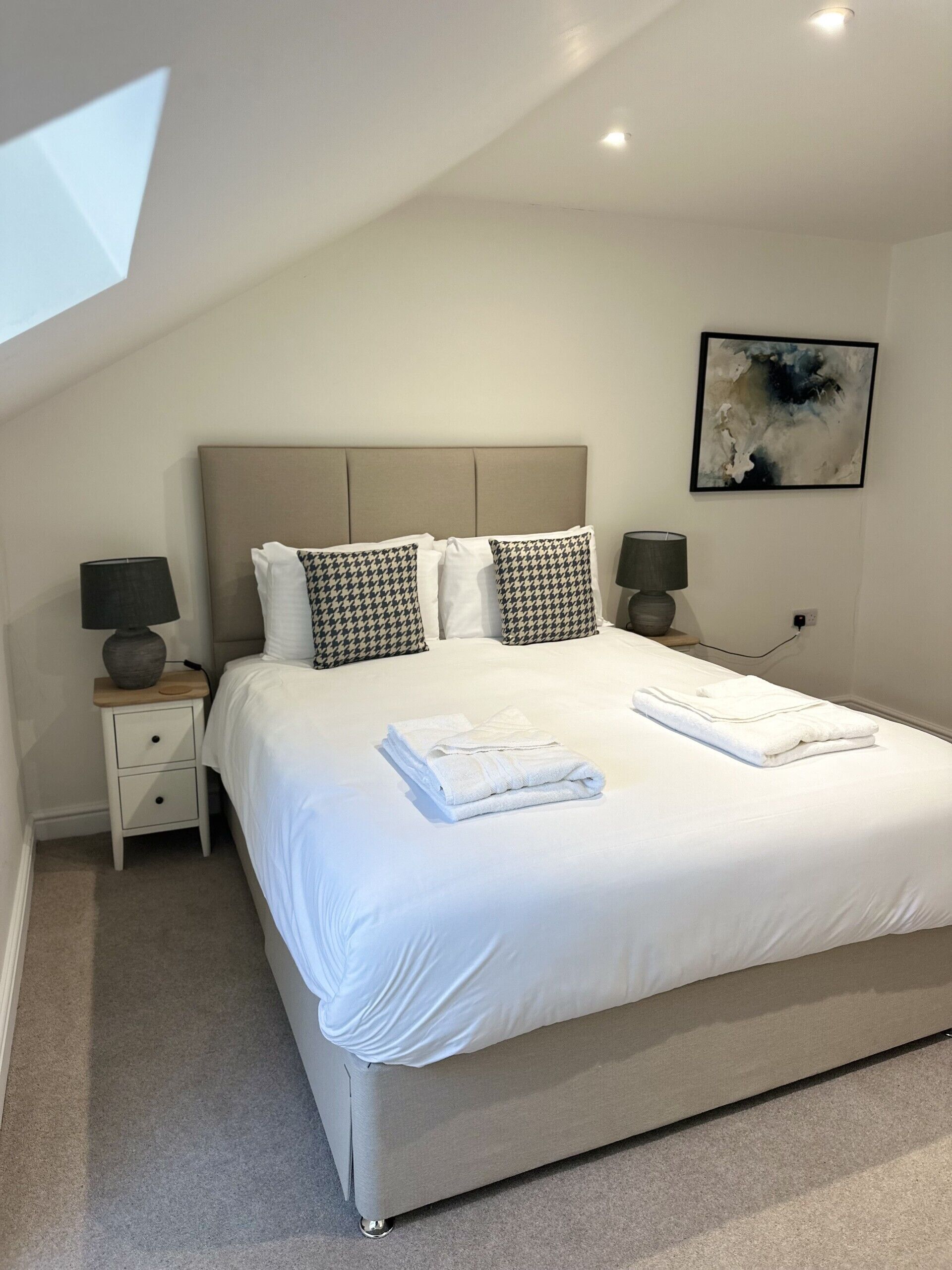 Un lit bien fait, avec du linge de lit blanc et deux coussins à carreaux, une table de chevet avec une lampe, et une peinture abstraite moderne accrochée au mur.