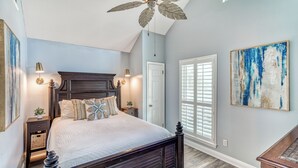 Une chambre avec un grand lit, un ventilateur de plafond, une fenêtre avec des stores et un tableau au mur.