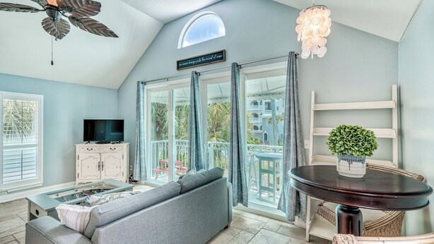 A spacious living room with a sofa, a foosball table, a TV, and a balcony with a view of the outdoors. - Adorable Upstairs 2 BR Nantucket 6AB (Destin)