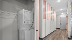 A laundry room with a stacked washer and dryer, a metal rack, and a door leading to a hallway with lockers.