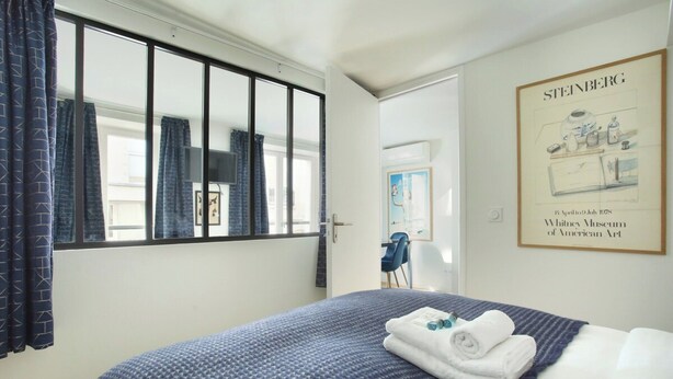 Una camera d'albergo con un letto, una finestra con tende, un quadro appeso al muro e una sedia nell'angolo.