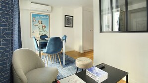 Un soggiorno con un divano, un tavolo da pranzo con sedie, un poggiapiedi e un quadro incorniciato, oltre a un poster appeso al muro.