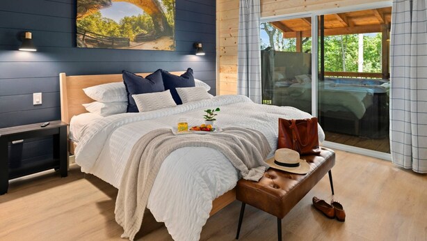 A bedroom with a large bed, a nightstand, a bedside table with a hat and shoes, and a painting on the wall. - New 4-King Cabin w/ Cave View, Hot Tub, Fire Pit (Frenchburg)