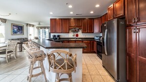 A spacious kitchen with wooden cabinets, a central island with bar stools, a stainless steel refrigerator, and a dining area with a sofa and chairs. - Dog‑Friendly (Only 1) Bayside Condo w/ Pool & Gym (Selbyville)
