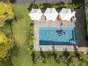 Vue aérienne d’une piscine avec un flotteur gonflable bleu, entourée d’une terrasse carrelée et de parasols blancs.