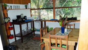 A kitchen with wooden cabinets, a dining table with chairs, a window with a view of greenery, and a trash bin. - Paradiselodge - Romantic Hideaway, next to Nauyaca (Platanillo de Baru)