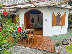 A small, single-story house with a wooden deck, a covered porch, and a garden area with potted plants and a red flower.