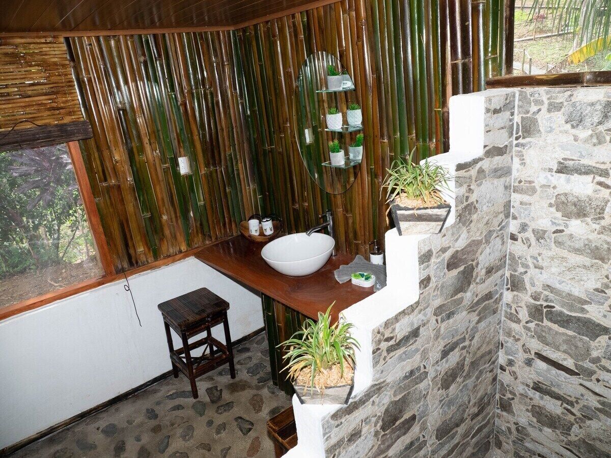 Un baño con pared de piedra, encimera de madera, lavabo blanco y espejo. Hay dos plantas en macetas y un taburete pequeño.