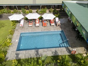 An outdoor swimming pool with lounge chairs and umbrellas, surrounded by greenery and a building with a green roof.