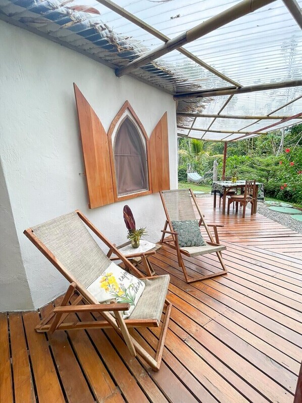 A wooden deck with a covered seating area, a table, and a view of a garden.