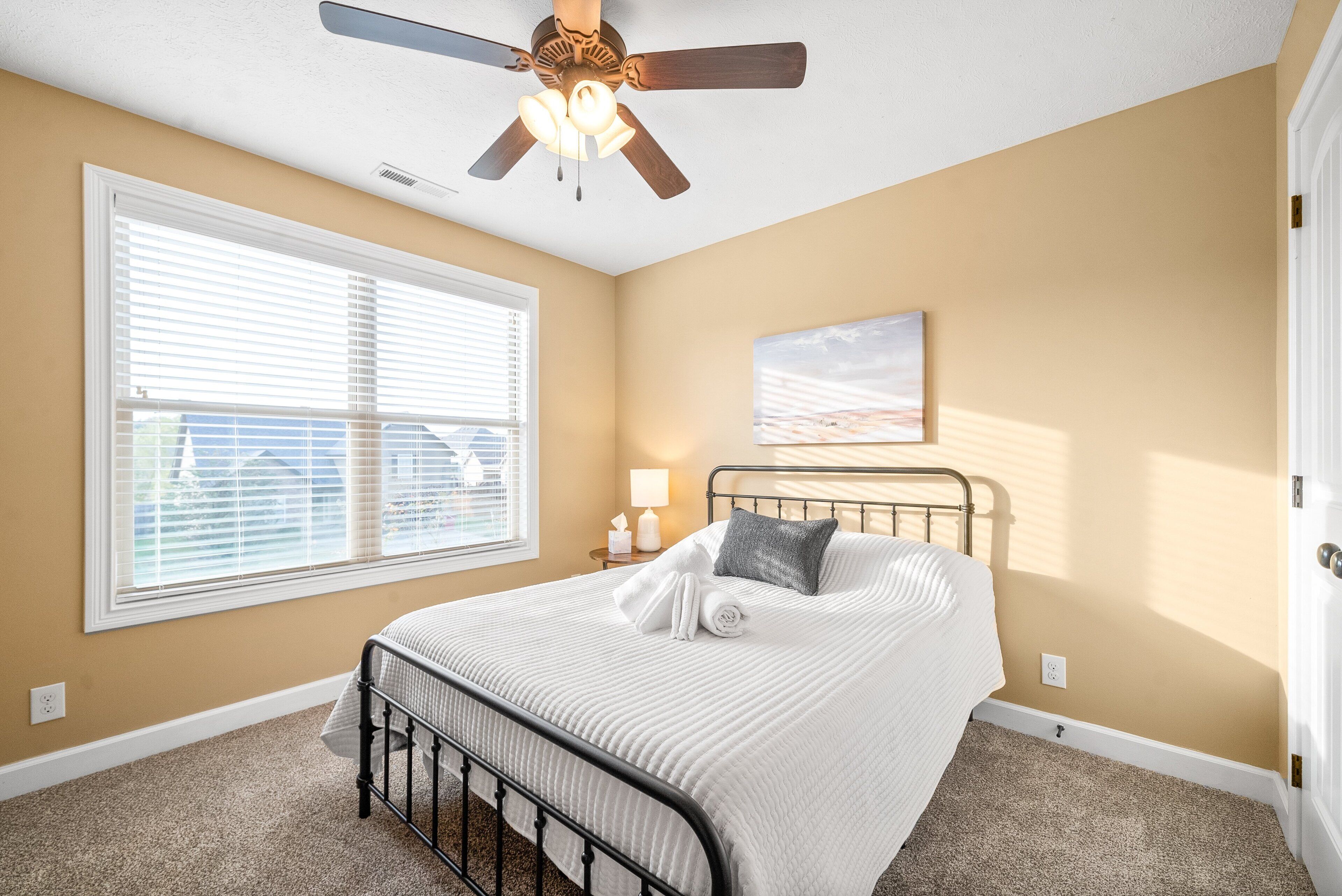 Une chambre à coucher avec un lit, un ventilateur de plafond, une fenêtre avec des stores et un tableau au mur.