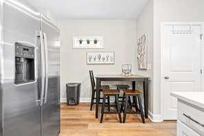 Une cuisine moderne équipée d’un réfrigérateur en acier inoxydable, d’une table à manger avec quatre chaises, d’une poubelle et d’une fenêtre agrémentée de plantes en pot.