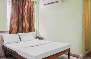 A single bed with white linens in a room with light green walls, a window with patterned curtains, and an air conditioning unit on the wall.