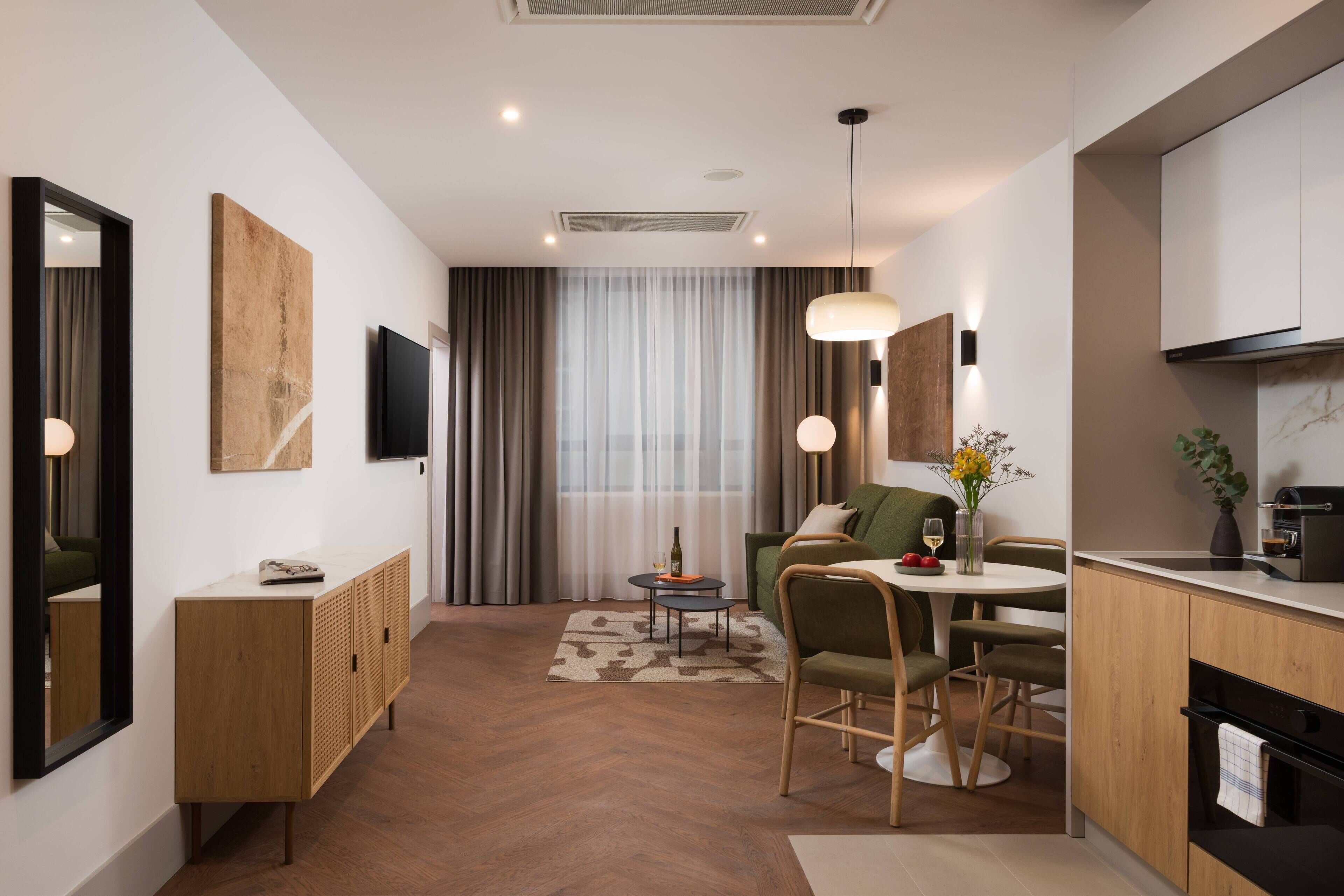 A modern hotel room with a wooden floor, a dining area with a table and chairs, a kitchen with a stove and sink, and a large window with curtains.