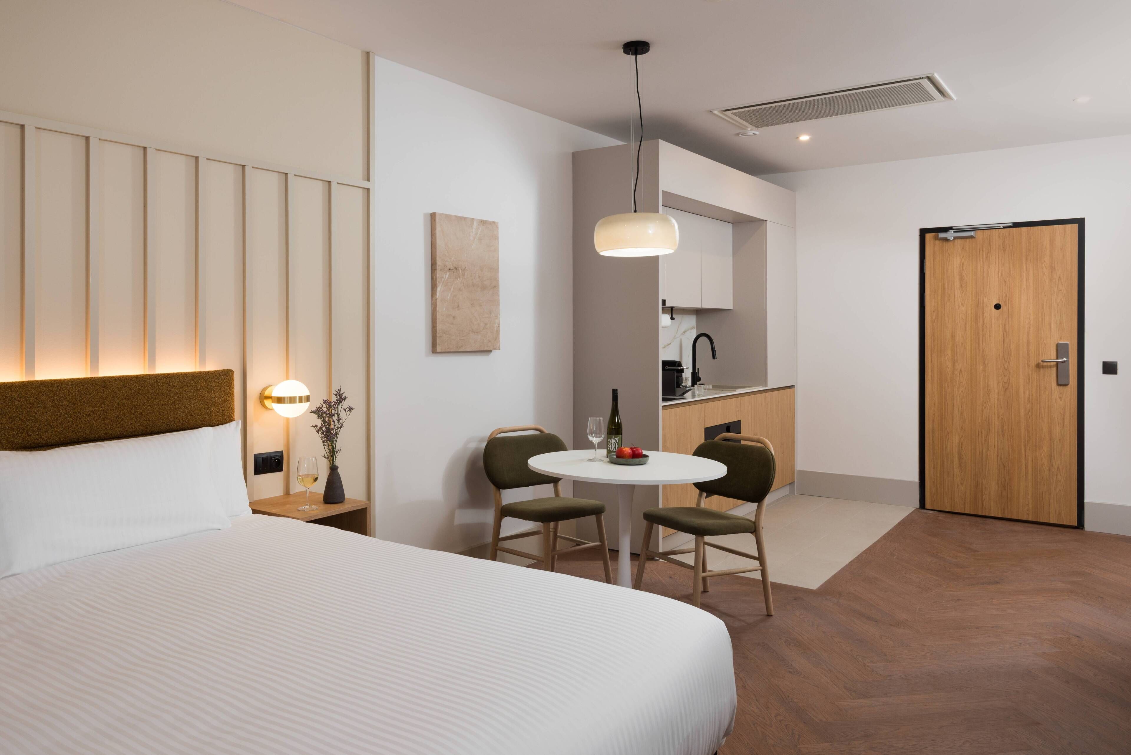Habitación de hotel con una cama grande, una pequeña mesa redonda con sillas, una kitchenette y una puerta de madera.
