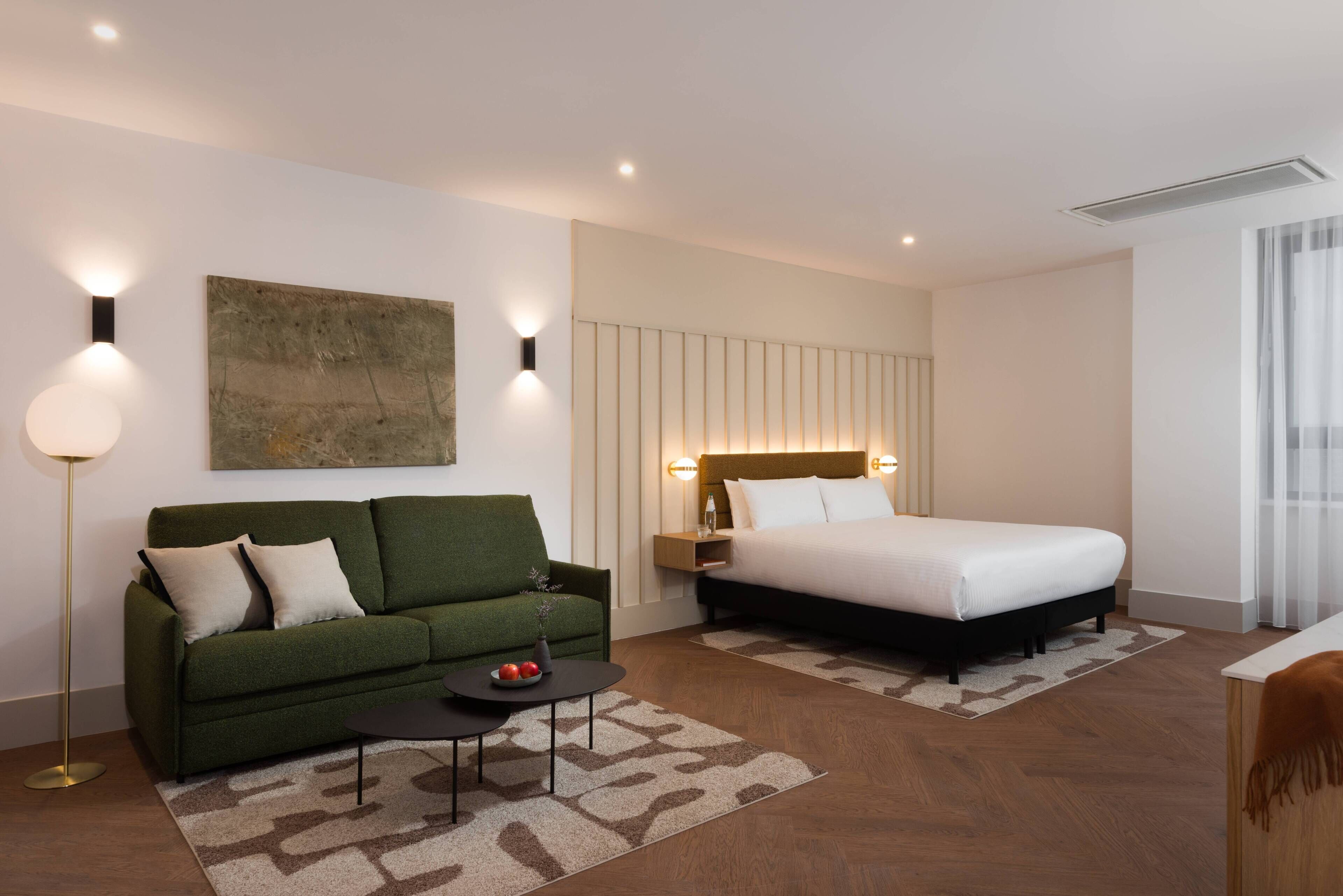 A modern hotel room with a green sofa, a bed with a wooden headboard, a small table with a vase, and a painting on the wall.