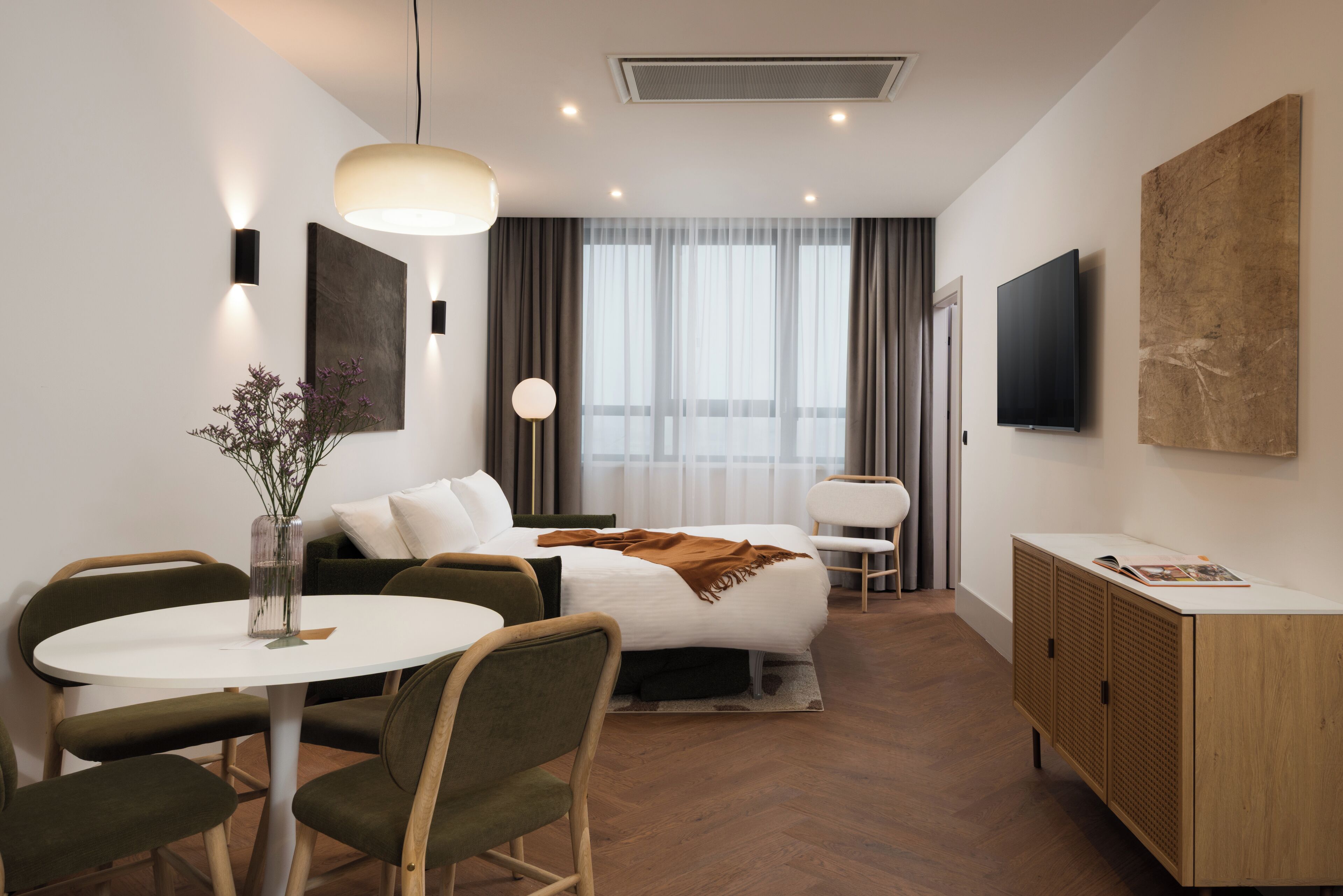 A modern hotel room with a bed, a round dining table, two chairs, a small side table, a TV, a cabinet, and a large window with curtains.
