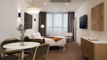A modern hotel room with a bed, a round dining table, two chairs, a small side table, a TV, a cabinet, and a large window with curtains.