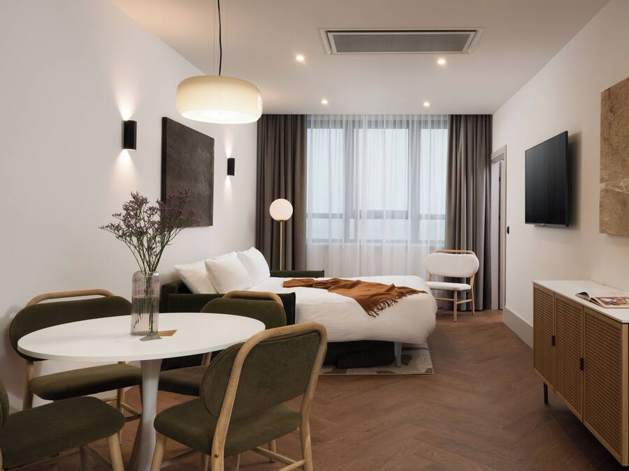 A modern hotel room with a bed, a round dining table, two chairs, a small side table, a TV, a cabinet, and a large window with curtains.