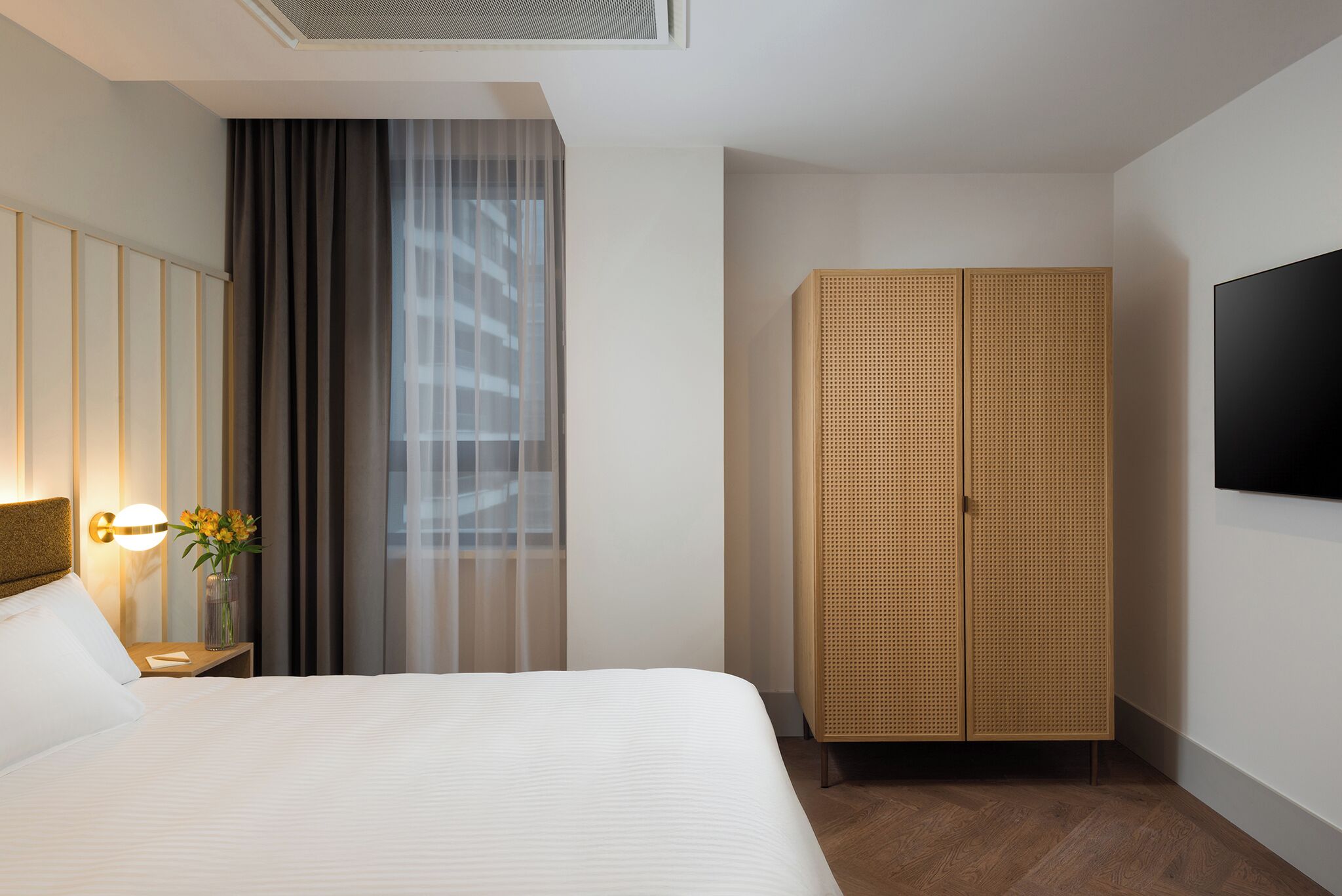 Une chambre d’hôtel avec un lit, une table de chevet avec une lampe, un vase de fleurs, une armoire et un téléviseur fixé au mur.