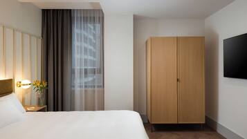 Une chambre d’hôtel avec un lit, une table de chevet avec une lampe, un vase de fleurs, une armoire et un téléviseur fixé au mur.
