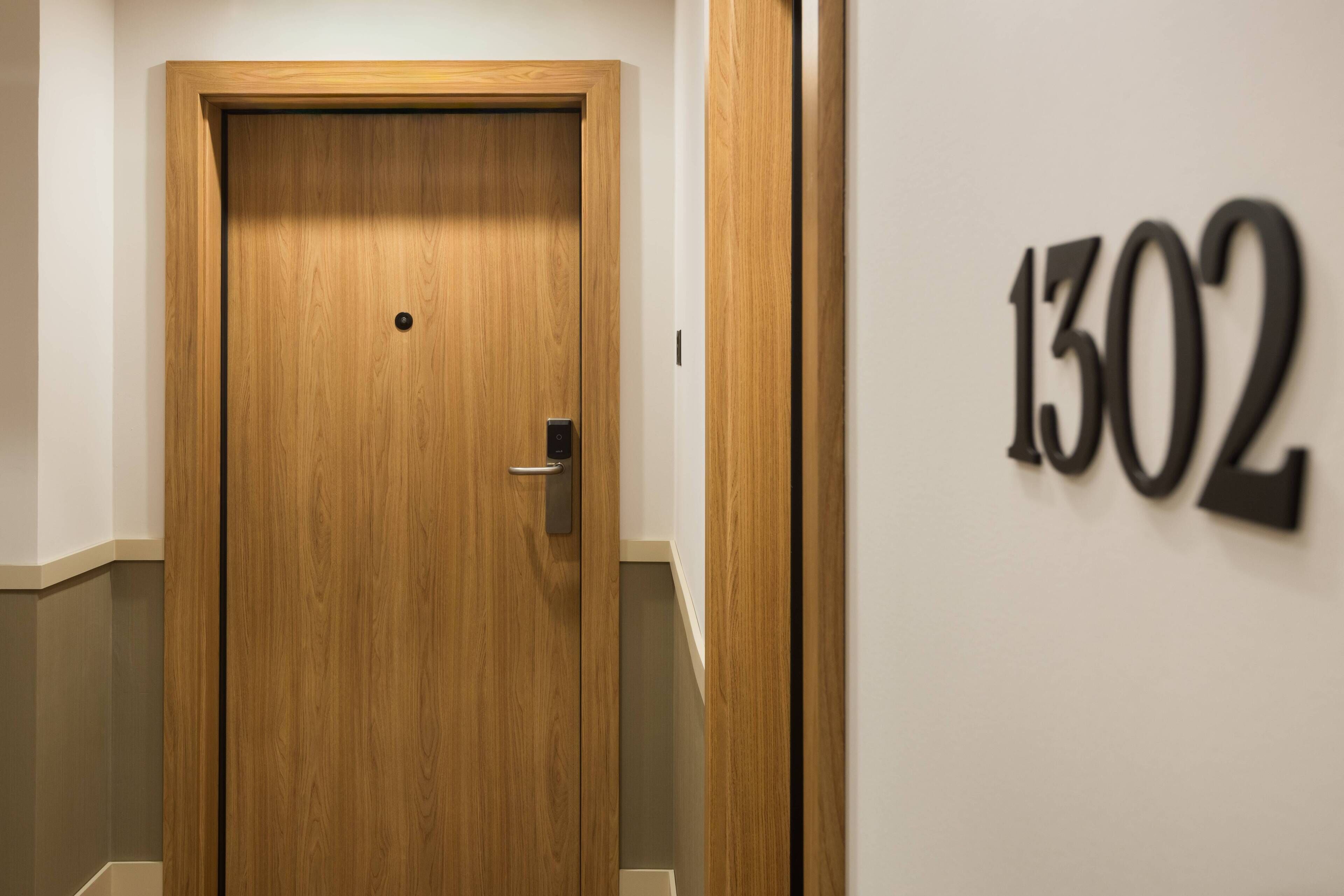 Une porte en bois, dotée d’une poignée et d’un petit trou au centre. Une plaque fixée au mur, portant le numéro « 1302 », est visible à droite de la porte.