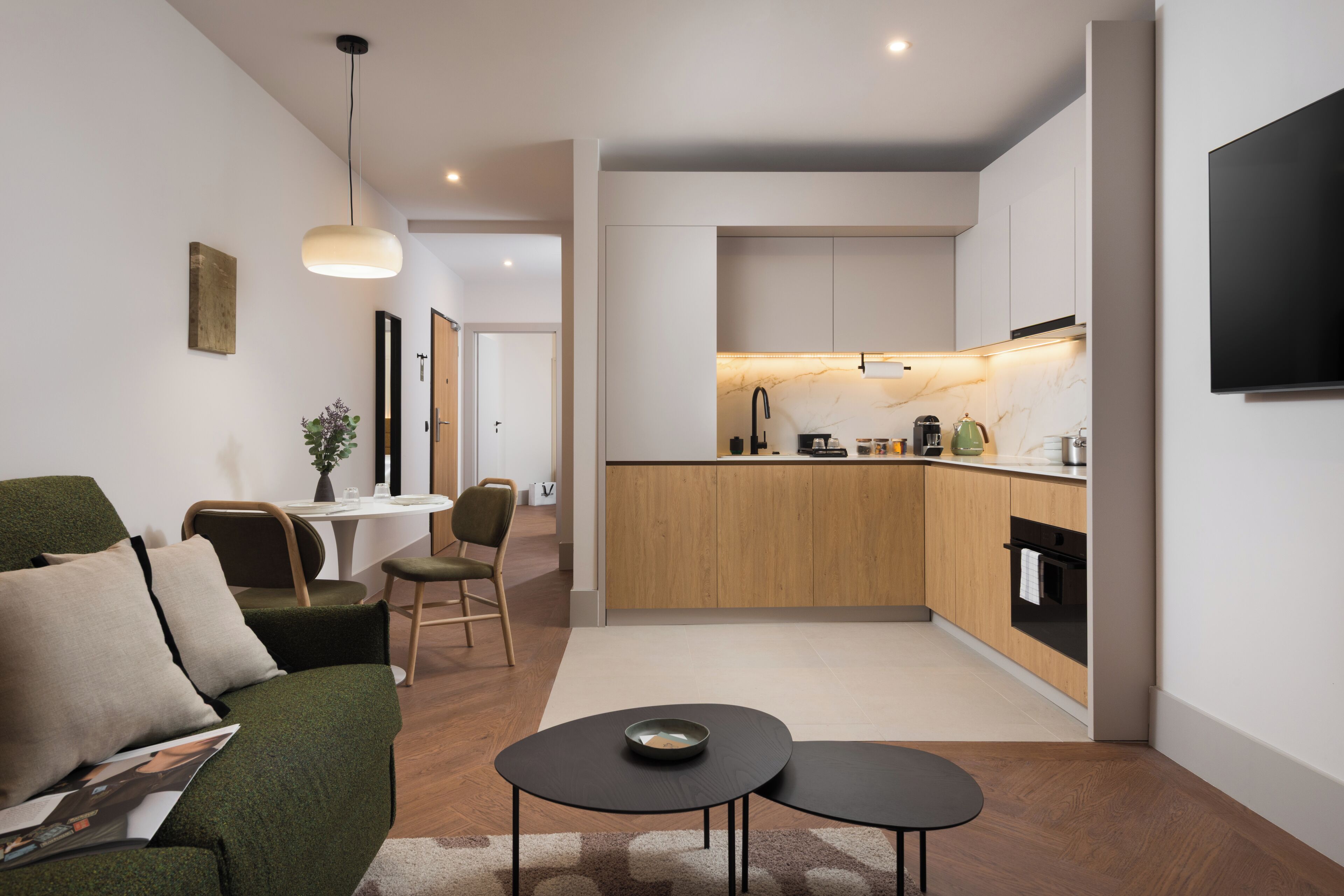 A modern kitchen with wooden cabinets, a built-in oven, and a dining area with a green sofa and two black tables.