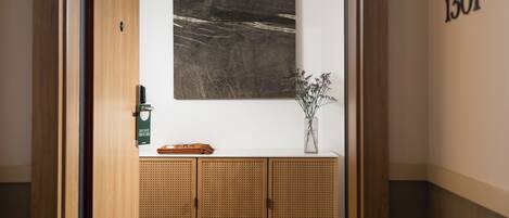 A hallway with a wooden door, a white wall, a number '1301' on the right, a woven cabinet with a vase of flowers, and a black and white abstract painting above the cabinet.
