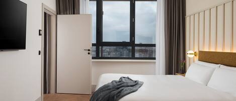 Habitación de hotel con cama, televisor de pantalla plana, ventana con cortinas y vista a la ciudad.