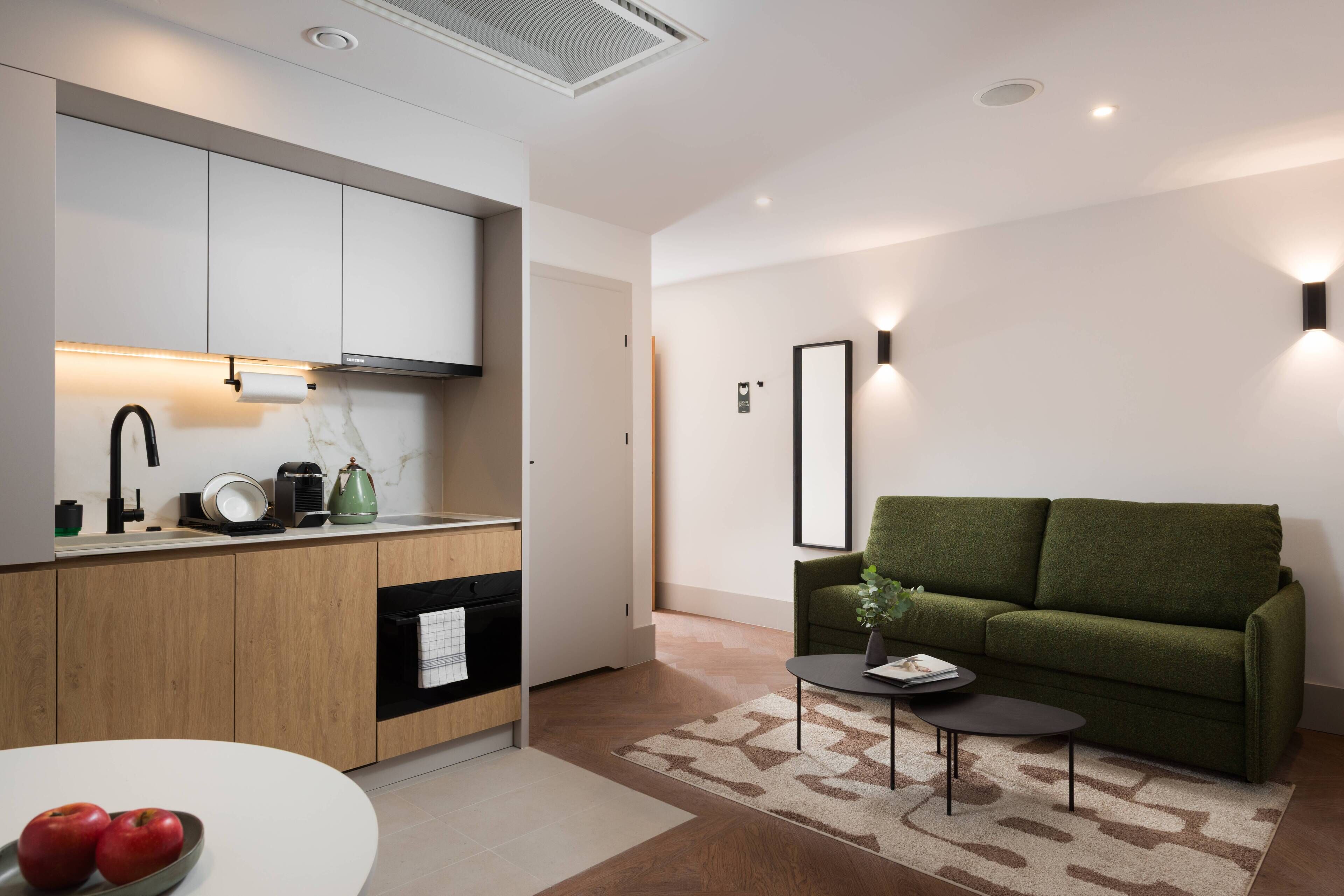 A modern kitchen with wooden cabinets, a built-in oven, and a countertop with a kettle and coffee maker. There is a green sofa and a small table with a book and a vase in the living area.