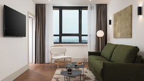 A modern living room with a flat-screen TV, a green sofa, a white armchair, a small coffee table with a croissant, and a view through a large window. - master Wola (Warsaw)