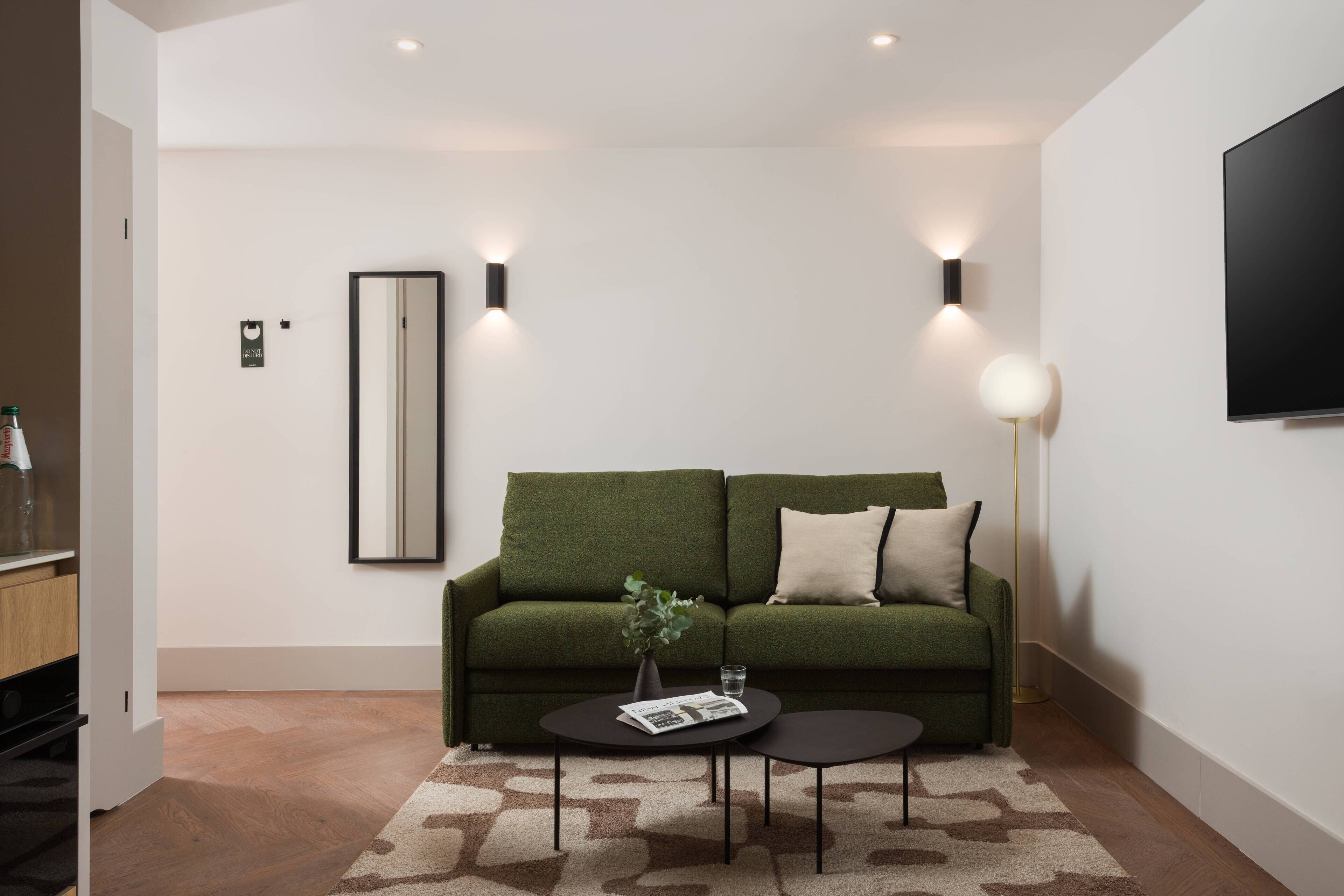 A modern living room with a green sofa, two black coffee tables, a floor lamp, and a flat-screen TV mounted on the wall.
