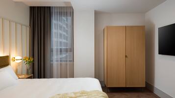 Quarto de hotel com uma cama grande, um guarda-roupa de madeira, uma mesa de cabeceira com abajur e uma televisão na parede.