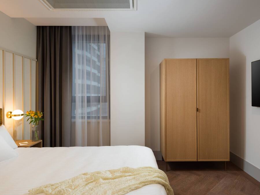 Quarto de hotel com uma cama grande, um guarda-roupa de madeira, uma mesa de cabeceira com abajur e uma televisão na parede.
