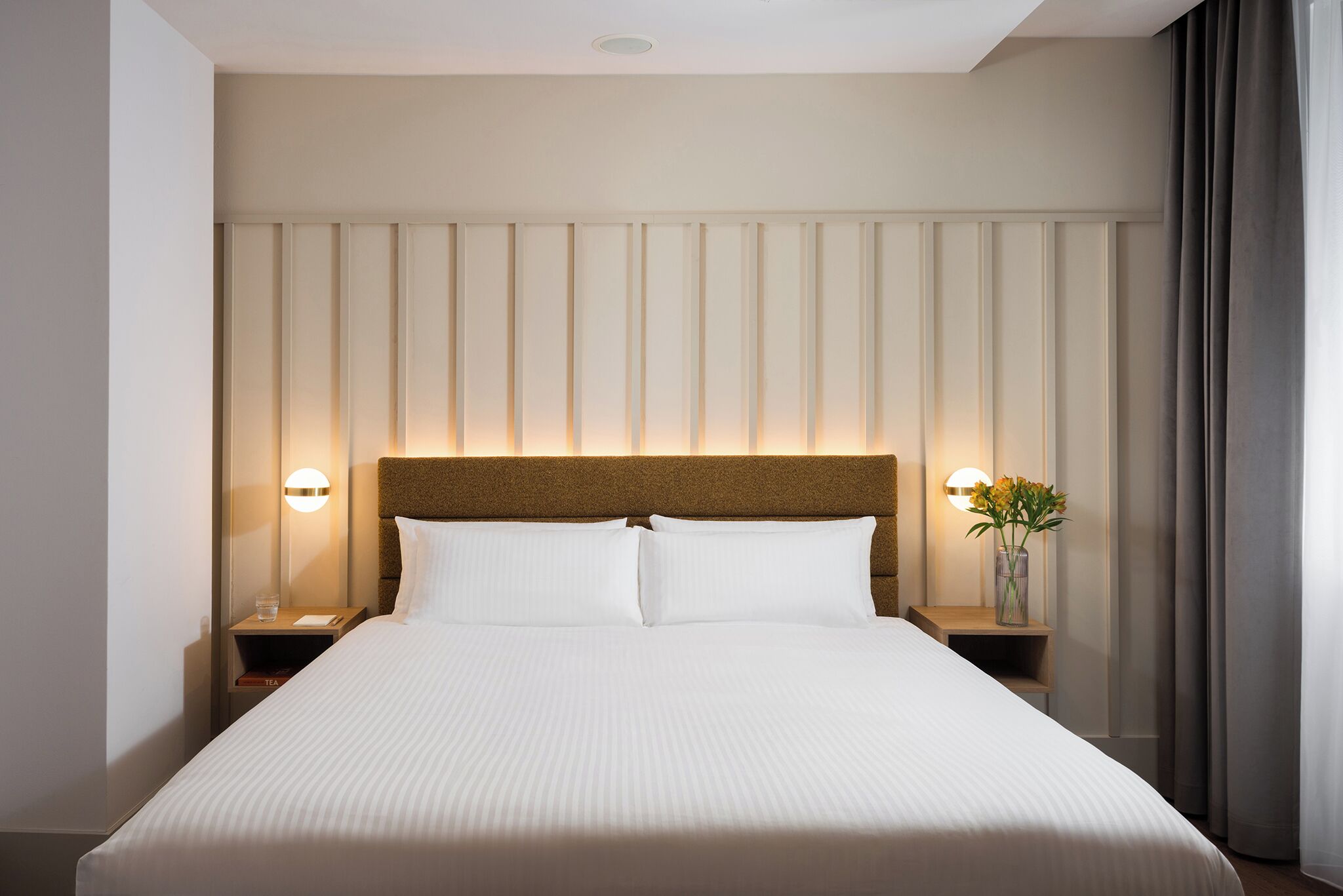 A hotel room with a large bed, two bedside tables, a nightstand, a vase with flowers, and a wall-mounted light fixture.