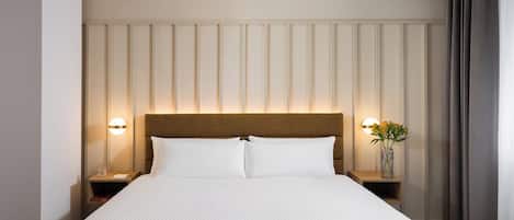 A hotel room with a large bed, two bedside tables, a nightstand, a vase with flowers, and a wall-mounted light fixture.