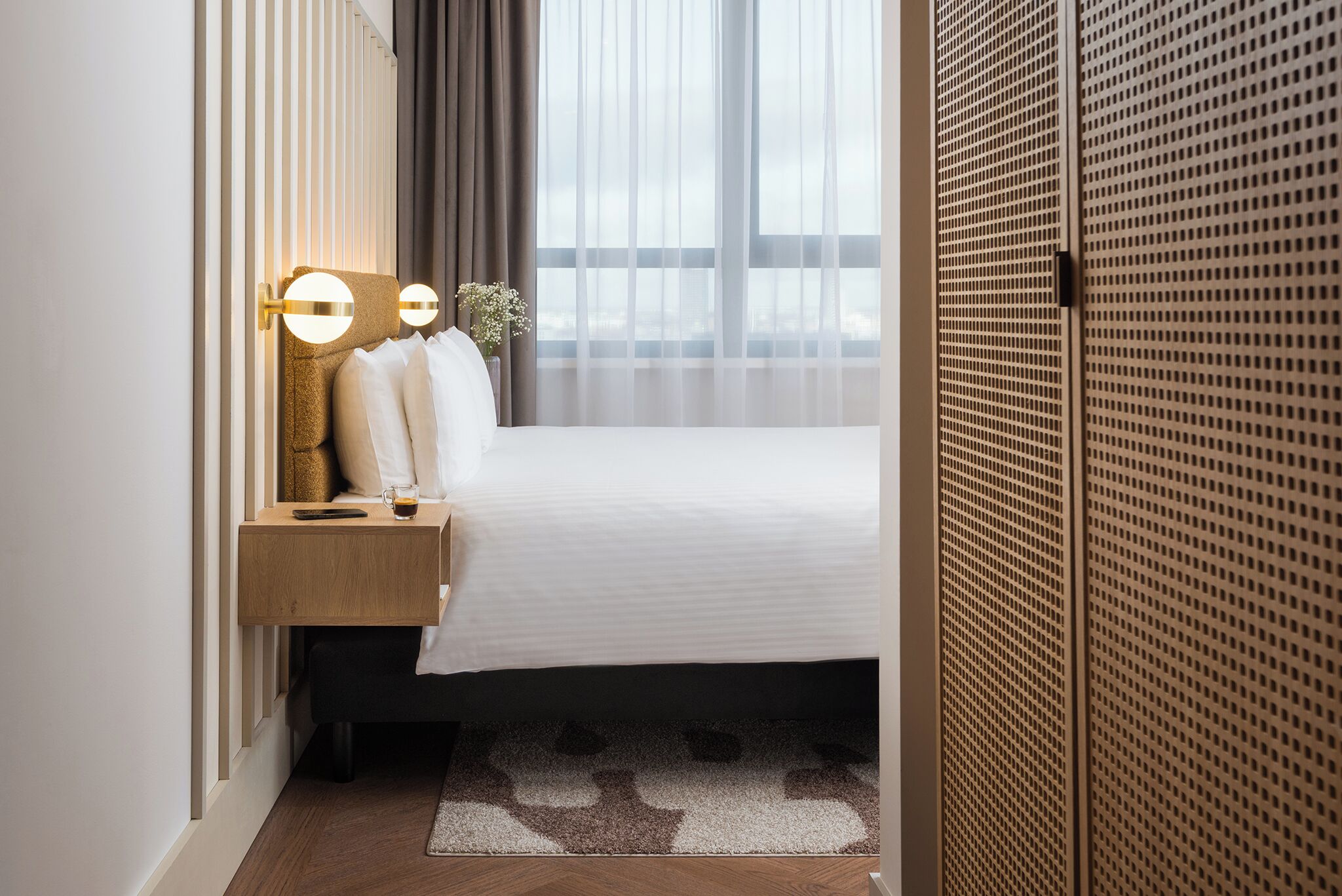 Quarto de hotel bem arrumado, com cama, mesa de cabeceira, abajur e vista para o exterior através de cortinas translúcidas.