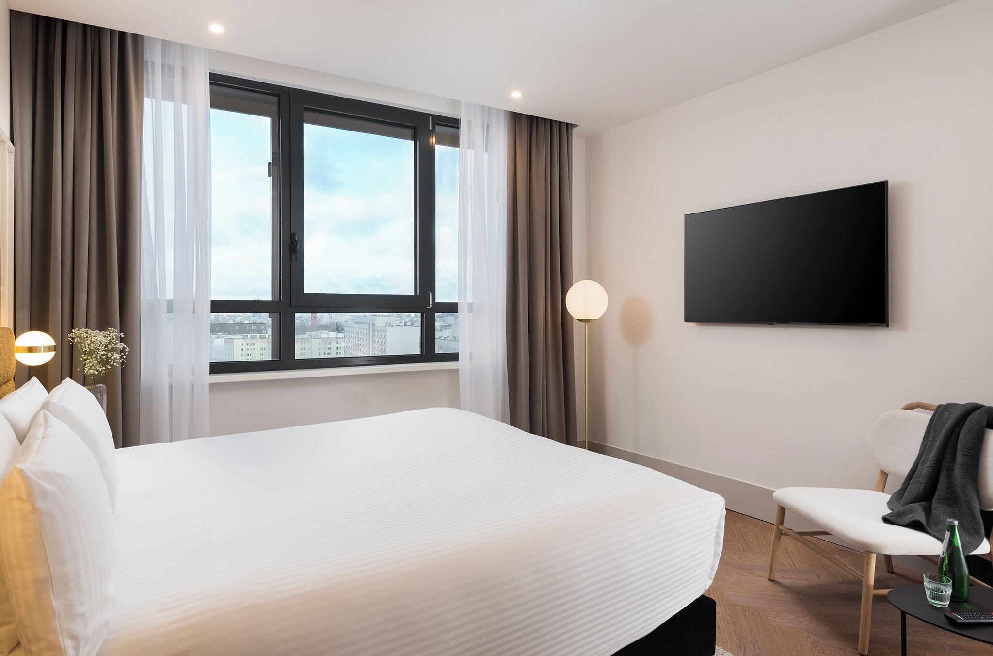 A hotel room with a large bed, a TV mounted on the wall, a chair, and a window with curtains.