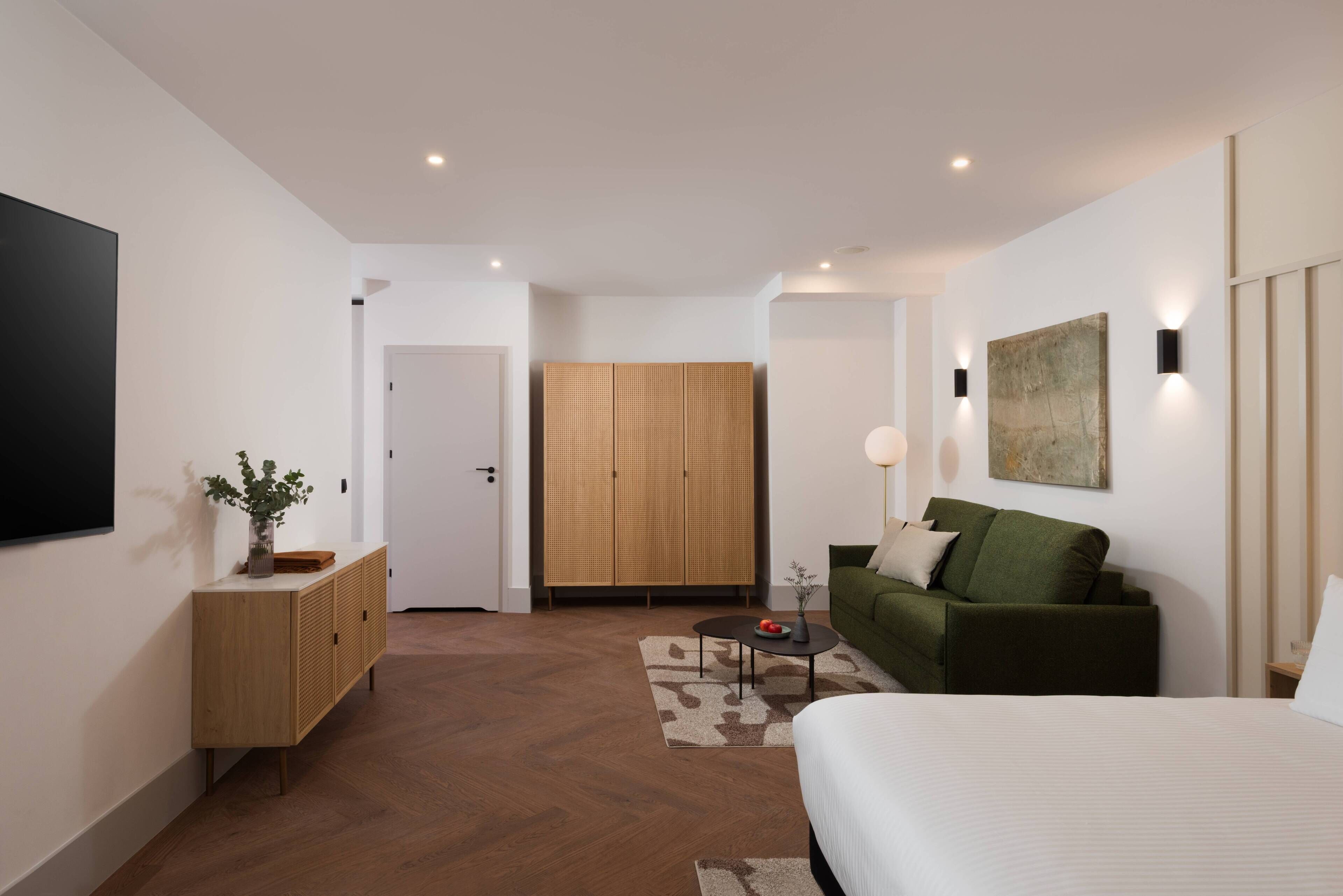 Una habitación de hotel moderna con una cama grande, un sofá verde, un armario de madera y un televisor de pantalla plana montado en la pared.