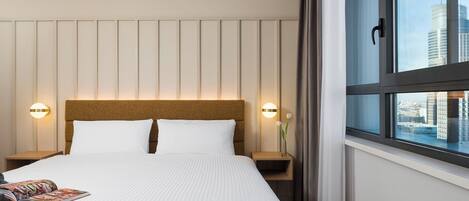 A hotel room with a large bed, two bedside tables, a nightstand, a window with a view of the city, and a headboard with vertical paneling.