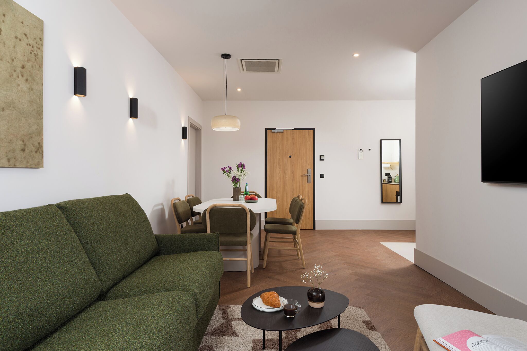 Una sala moderna con un sofá verde, un comedor con mesa y sillas blancas, una puerta de madera y un televisor montado.